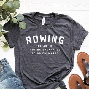 Peut inclure: Un t-shirt gris foncé avec le texte "ROWING THE ART OF MOVING BACKWARDS TO GO FORWARDS" en lettres blanches. Le t-shirt est posé sur une surface blanche avec une paire de sandales marrons et un jean bleu.
