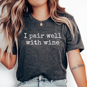 以下が含まれることがあります： 白い文字で「I pair well with wine」とプリントされた濃いグレーのTシャツ。モデルは薄いブルーのジーンズと白いスニーカーを合わせています。