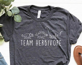 Op de afbeelding: Een donkergrijs T-shirt met een witte afbeelding van drie dinosaurussen en de tekst "TEAM HERBIVORE". Het shirt ligt op een witte ondergrond met een paar bruine sandalen en een blauwe spijkerbroek.