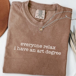 May include: Espresso-colored t-shirt with the text "everyone relax i have an art degree" in white. The shirt has a crew neck and a small tag on the collar. A silver necklace with a pendant is draped across the shirt.