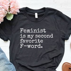 May include: Black t-shirt with white text that reads "Feminist is my second favorite F-word."