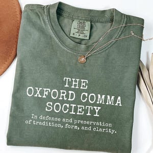 May include: A moss green t-shirt with the text "THE OXFORD COMMA SOCIETY" in white. The shirt also has the phrase "In defense and preservation of tradition, form, and clarity." A gold necklace and a brown hat are also in the image.