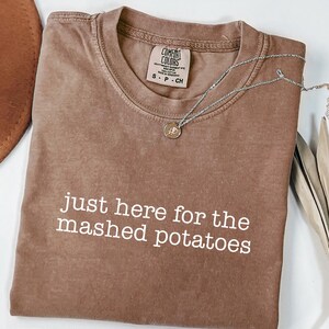 May include: Espresso-coloured t-shirt with the text "just here for the mashed potatoes" in white. The shirt has a crew neck and a Comfort Colors tag. A silver necklace with a pendant is draped across the shirt.