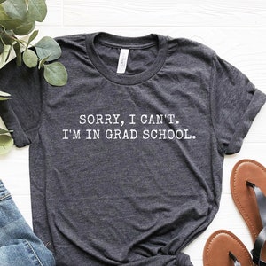 May include: A dark gray t-shirt with the text "SORRY, I CAN'T. I'M IN GRAD SCHOOL." printed in white.