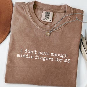 May include: Espresso-colored t-shirt with the text "i don't have enough middle fingers for MS" in white. A silver necklace with a small pendant is draped across the shirt. The shirt is folded, and a brown hat is partially visible.