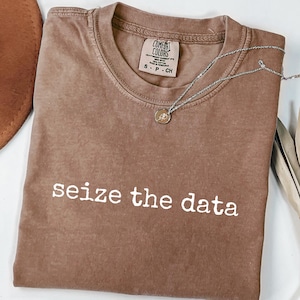 May include: Espresso-coloured t-shirt with the phrase "seize the data" in white lettering. The shirt has a crew neck and short sleeves. A silver necklace with a small pendant is draped across the shirt. The shirt is made by Comfort Colors.
