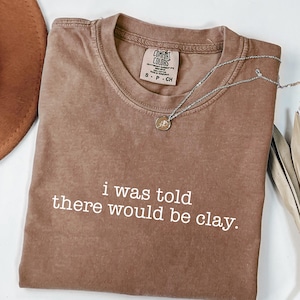 May include: Espresso-coloured t-shirt with the text "i was told there would be clay" in white. The shirt has a crew neck and a small tag near the collar. A silver necklace with a pendant is draped across the shirt.