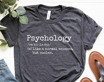 May include: A dark gray t-shirt with the word "Psychology" defined as "Like a normal science, but cooler." The shirt is laid flat on a white surface with a pair of brown sandals and blue jeans.