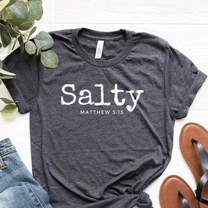 May include: A dark gray t-shirt with the word "Salty" printed in white with the text "Matthew 5:13" underneath. The shirt is laid out on a white wooden surface with a pair of blue jeans and brown sandals.