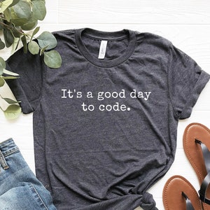 May include: A dark heather grey t-shirt with the text "It's a good day to code." printed in white.