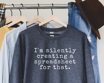 May include: A dark grey sweatshirt with the text "I'm silently creating a spreadsheet for that" in white. The sweatshirt is hanging on a clothes rail with other shirts and a brown hat.