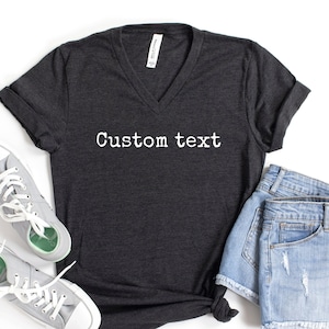 May include: A dark gray V-neck t-shirt with the text "Custom text" printed in white on the front. The shirt is paired with denim shorts and gray sneakers.