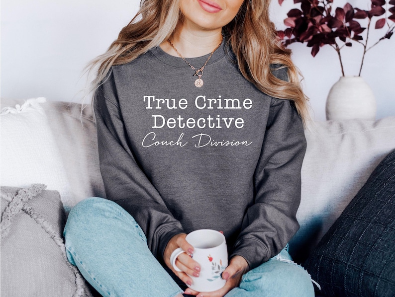 K&ouml;nnte beinhalten: Ein grauer Pullover mit dem Text "True Crime Detective Couch Division" in wei&szlig;en Buchstaben. Der Pullover wird von einer Person getragen, die auf einem Sofa sitzt.