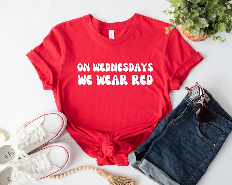 May include: Red t-shirt with white text that reads "On Wednesdays We Wear Red". The shirt is styled with denim shorts, white sneakers, and wooden beads.