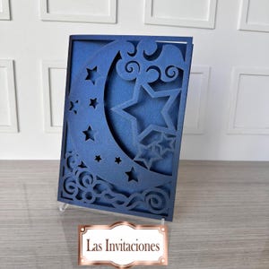 Puede incluir: Una tarjeta azul marino con un diseño celestial. La tarjeta presenta recortes de luna creciente, estrellas y nubes arremolinadas. El texto "Las Invitaciones" se muestra en una etiqueta dorada en la parte inferior. La tarjeta se muestra en un soporte transparente.