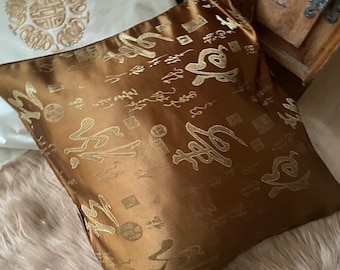 May include: A brown satin pillow with gold embroidered Chinese characters and symbols. The pillow is on a light brown fuzzy surface.