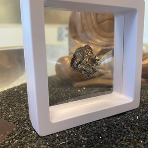 May include: A small, dark gray meteorite encased in a clear plastic square display case with a white frame. The case is sitting on a bed of black sand.