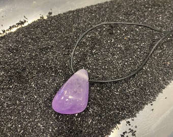 Puede incluir: Un colgante de amatista púrpura en un cordón de cuero negro. El colgante tiene forma de lágrima y está sobre un lecho de arena negra.