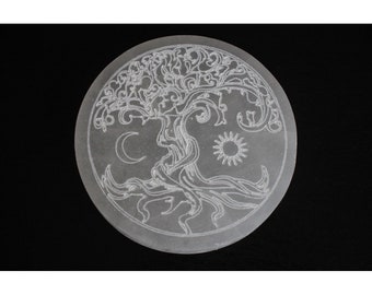 May include: A white, round, polished stone with a carved tree of life design. The tree has intricate branches and roots, and is surrounded by a crescent moon and a sun.