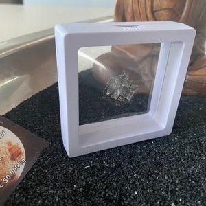 May include: A small, gray, metallic meteorite in a clear plastic display case with a white frame. The case is sitting on a bed of black sand.