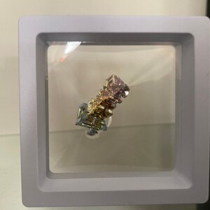 May include: A colorful bismuth crystal displayed in a clear acrylic cube with a gray frame. The crystal has a metallic sheen and iridescent colors, including purple, gold, and silver.