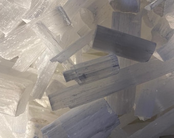 May include: A pile of white selenite crystal sticks. The sticks are various sizes and shapes, and some have a rough, textured surface.