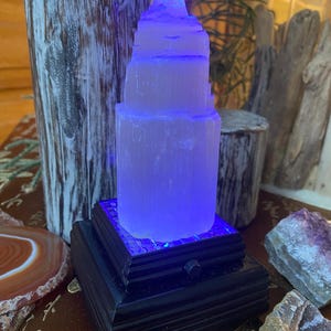 Könnte beinhalten: Ein weißer Selenit-Turm mit einer blauen Lichtbasis auf einem schwarzen Holzsockel. Der Turm besteht aus übereinander gestapelten Selenitkristallen und wird von unten durch ein blaues LED-Licht beleuchtet. Der Sockel hat ein dekoratives Design und ist aus Holz.