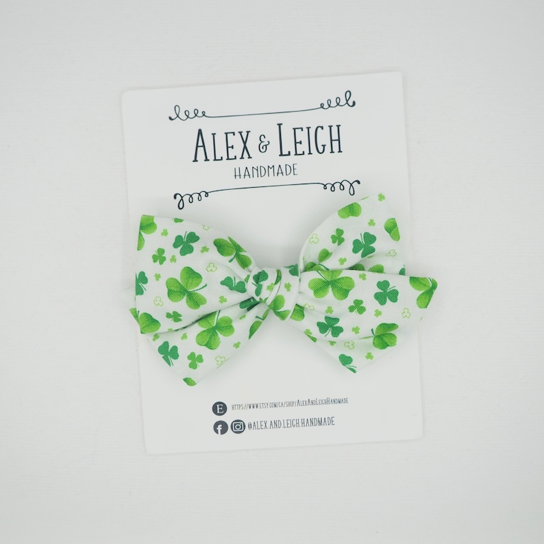 May include: A white fabric bow with a green shamrock pattern. The bow is handmade and has the text "Alex & Leigh Handmade" on a white card behind it.