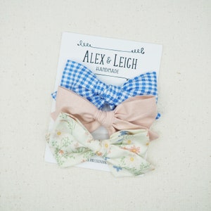 May include: Three handmade hair bows in blue gingham, blush pink, and a floral print. The bows are stacked on top of each other and are on a white background. The text "Alex & Leigh Handmade" is visible on the card behind the bows.