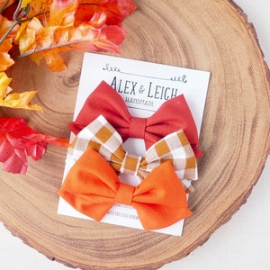 Pu&ograve; includere: Tre papillon in tessuto fatti a mano in arancione, rosso e a quadri marroni. I papillon sono disposti su una carta bianca con il testo "Alex & Leigh Handmade".