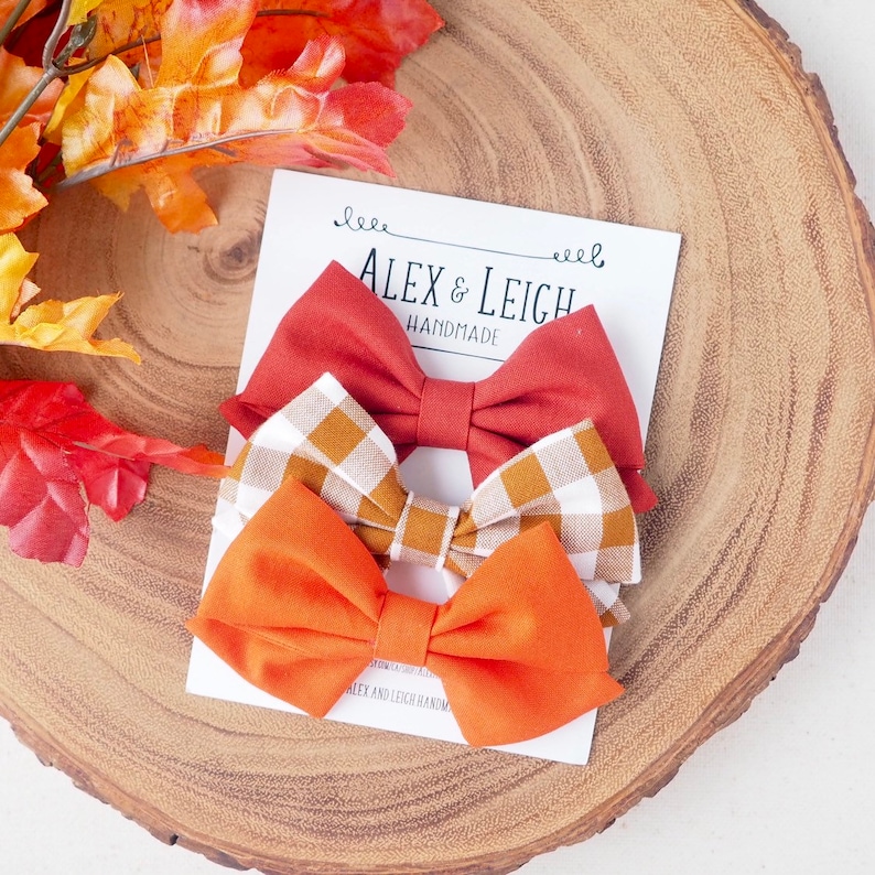 Pu&ograve; includere: Tre papillon in tessuto fatti a mano in arancione, rosso e a quadri marroni. I papillon sono disposti su una carta bianca con il testo "Alex & Leigh Handmade".
