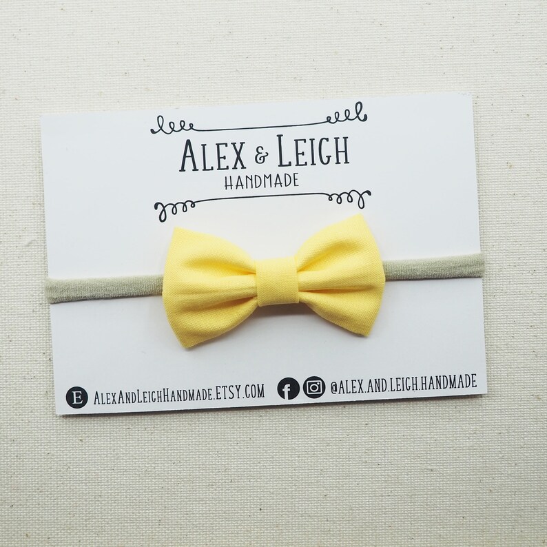 May include: A yellow bow headband with a nude elastic band. The headband is on a white card with the text "Alex & Leigh Handmade" printed on it.