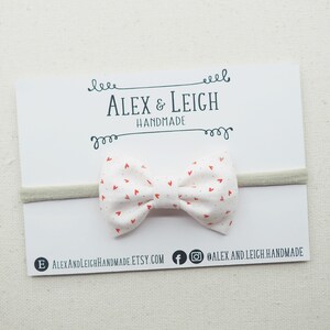 Peut inclure: Un bandeau blanc avec un n&oelig;ud en tissu imprim&eacute; de c&oelig;urs rouges. Le n&oelig;ud est nou&eacute; et poss&egrave;de une petite boucle &agrave; l'arri&egrave;re. Le bandeau est pos&eacute; sur une carte blanche avec le texte "Alex & Leigh Handmade" imprim&eacute; dessus.