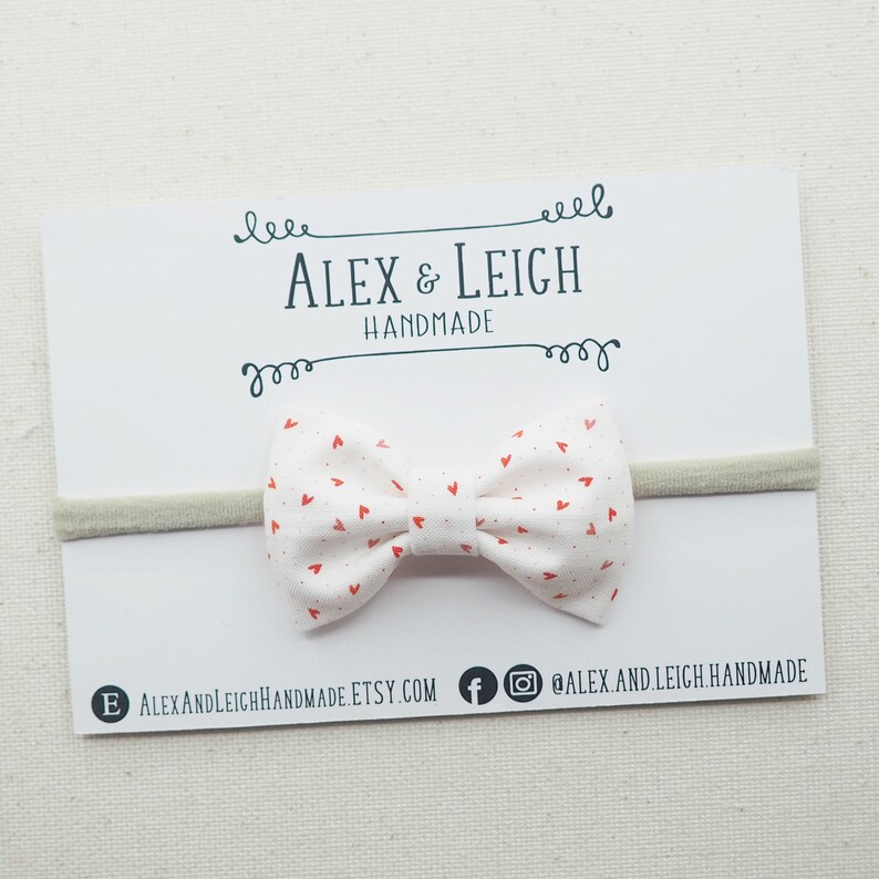Peut inclure: Un bandeau blanc avec un n&oelig;ud en tissu imprim&eacute; de c&oelig;urs rouges. Le n&oelig;ud est nou&eacute; et poss&egrave;de une petite boucle &agrave; l'arri&egrave;re. Le bandeau est pos&eacute; sur une carte blanche avec le texte "Alex & Leigh Handmade" imprim&eacute; dessus.