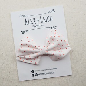 Peut inclure: Noeud en tissu blanc avec un motif de c&oelig;urs rouges. Le n&oelig;ud est fait &agrave; la main et est pr&eacute;sent&eacute; sur une carte blanche avec le texte "ALEX & LEIGH HANDMADE" et des informations sur les m&eacute;dias sociaux.