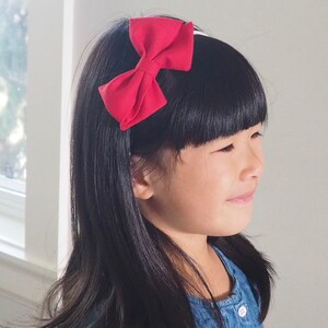 Pu&ograve; includere: Una bambina con lunghi capelli neri indossa un vestito di jeans blu con bottoni bianchi. Indossa una fascia per capelli in tessuto rosso con un fiocco.