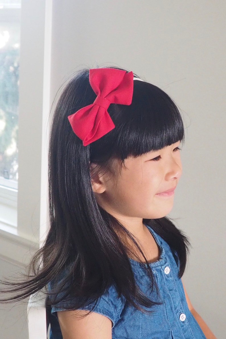 Pu&ograve; includere: Una bambina con lunghi capelli neri indossa un vestito di jeans blu con bottoni bianchi. Indossa una fascia per capelli in tessuto rosso con un fiocco.