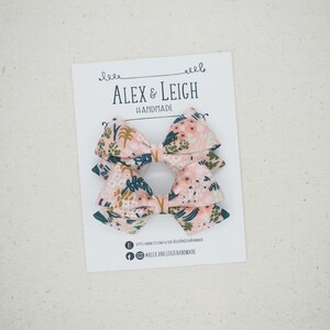 K&ouml;nnte beinhalten: Zwei rosa florale Haarb&auml;nder mit gr&uuml;nen Bl&auml;ttern und goldenen Akzenten. Die Schleifen befinden sich auf einer wei&szlig;en Karte mit dem Text "Alex & Leigh Handmade".