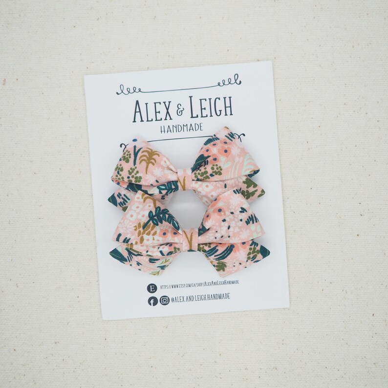 K&ouml;nnte beinhalten: Zwei rosa florale Haarb&auml;nder mit gr&uuml;nen Bl&auml;ttern und goldenen Akzenten. Die Schleifen befinden sich auf einer wei&szlig;en Karte mit dem Text "Alex & Leigh Handmade".
