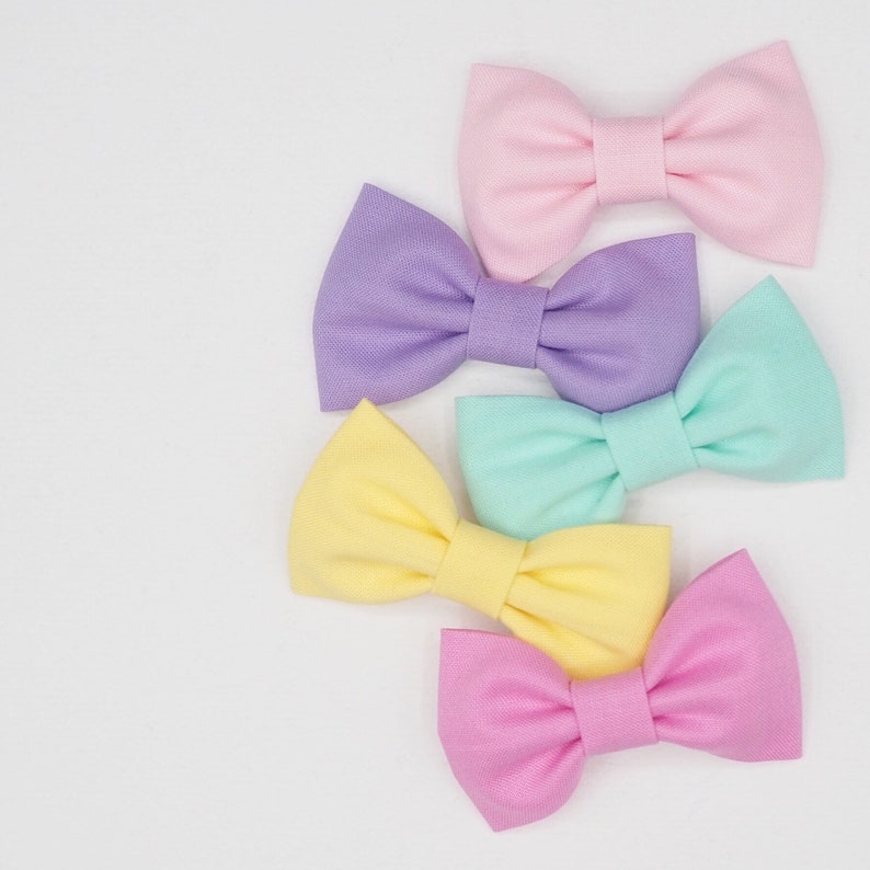 May include: Four pastel-colored fabric bow ties in pink, lavender, light blue, and yellow. The bows are arranged in a stack, with the pink bow on top and the yellow bow on the bottom.