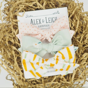 May include: Three handmade bow ties presented in a gift box. The bow ties are crafted from fabric with various patterns: pink with white crosses, light green with white flowers, and white with orange carrots. The box is filled with brown paper shreds.