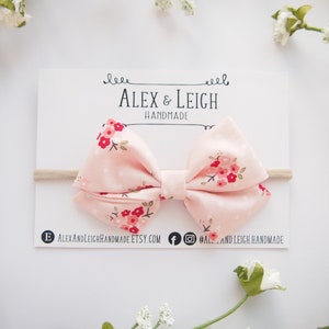 Puede incluir: Un lazo de tela rosa con estampado floral en tonos rojos y blancos. El lazo est&aacute; atado con una diadema el&aacute;stica de color nude. El lazo est&aacute; hecho a mano y se muestra en una tarjeta blanca con el texto "Alex & Leigh Handmade".
