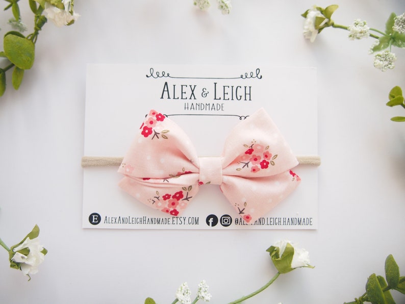 Puede incluir: Un lazo de tela rosa con estampado floral en tonos rojos y blancos. El lazo est&aacute; atado con una diadema el&aacute;stica de color nude. El lazo est&aacute; hecho a mano y se muestra en una tarjeta blanca con el texto "Alex & Leigh Handmade".