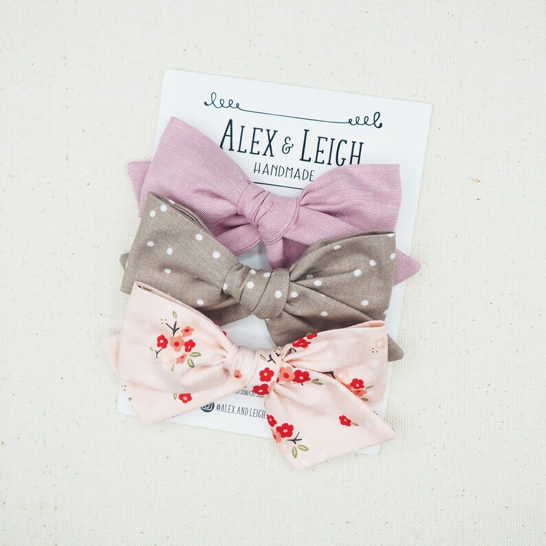 Puede incluir: Tres lazos de pelo de tela. El lazo superior es de color rosa s&oacute;lido, el lazo del medio es marr&oacute;n con lunares blancos y el lazo inferior es rosa con estampado floral.