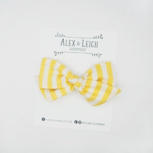 May include: Yellow and white striped fabric bow with a white card behind it. The card has the text "Alex & Leigh Handmade" on it.