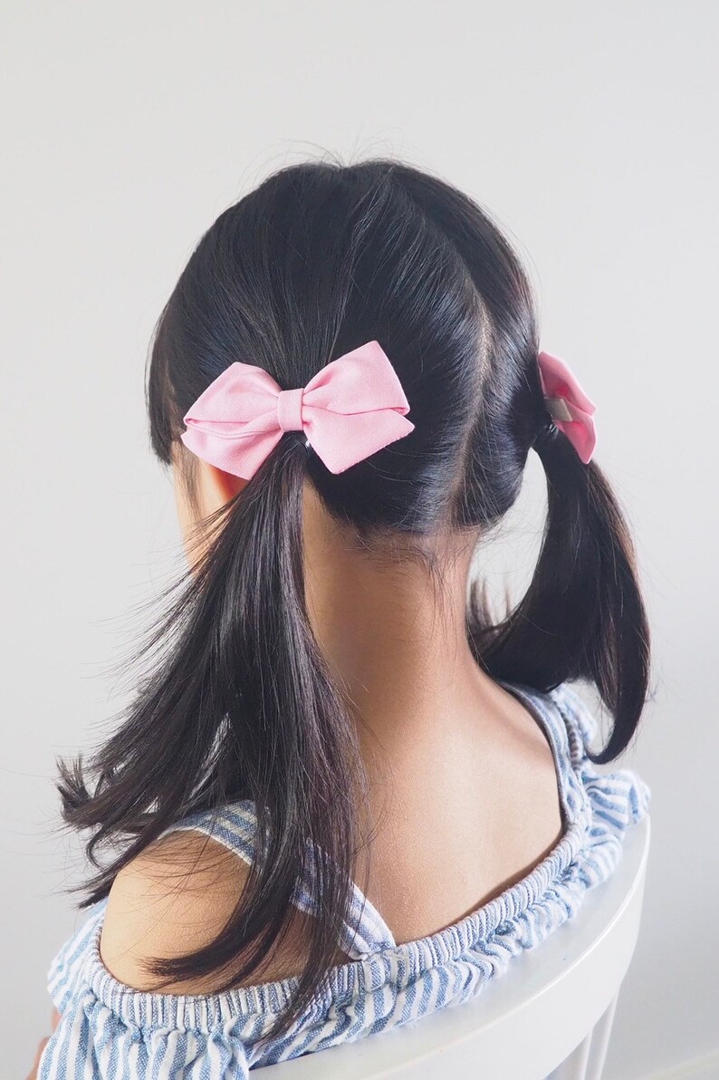 Pu&ograve; includere: Una bambina con lunghi capelli neri che indossa una camicetta blu e bianca a spalle scoperte con due fiocchi rosa nei capelli.