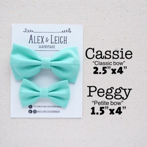 May include: Two handmade fabric bow ties in a light turquoise color. The larger bow tie is labeled "Cassie "Classic bow" 2.5"x4"" and the smaller bow tie is labeled "Peggy "Petite bow" 1.5"x4"".