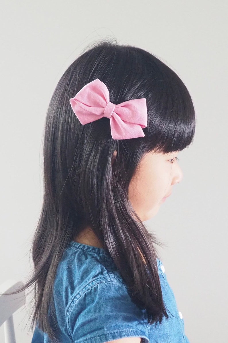 Pu&ograve; includere: Una bambina con lunghi capelli neri che indossa una camicia di jeans blu con un fiocco di stoffa rosa nei capelli.