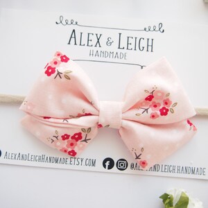 Puede incluir: Un lazo de tela rosa con estampado floral en tonos rojos y blancos. El lazo est&aacute; unido a una diadema el&aacute;stica de color nude.