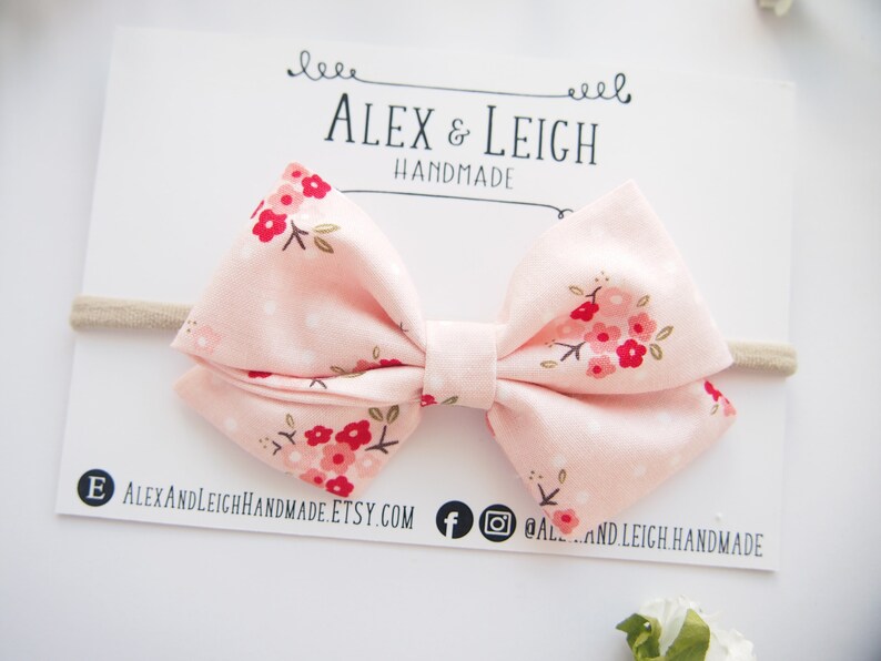 Puede incluir: Un lazo de tela rosa con estampado floral en tonos rojos y blancos. El lazo est&aacute; unido a una diadema el&aacute;stica de color nude.